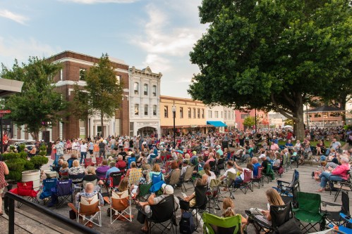 A large crowd gathers in a park for an outdoor event, seated in chairs with buildings and trees in the background.