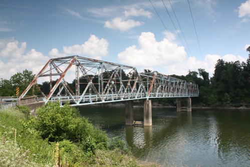 A metal truss bridge spans a calm river, surrounded by greenery and under a partly cloudy sky.