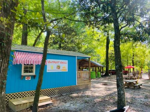 A colorful food stand named "Outpost Grill" surrounded by trees and picnic tables in a natural setting.