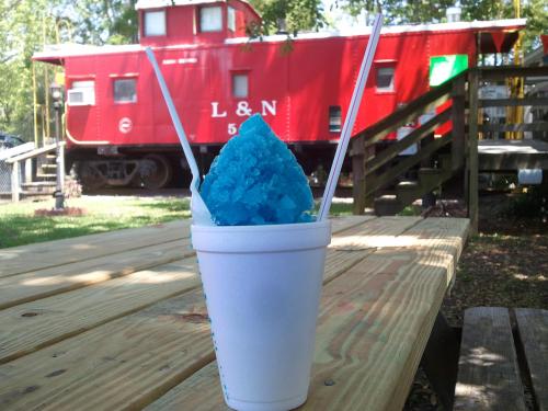 A blue snow cone in a white cup sits on a wooden table, with a red train caboose in the background.