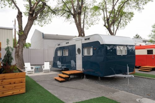 A vintage blue and white trailer parked on a gravel area, surrounded by trees and outdoor seating.