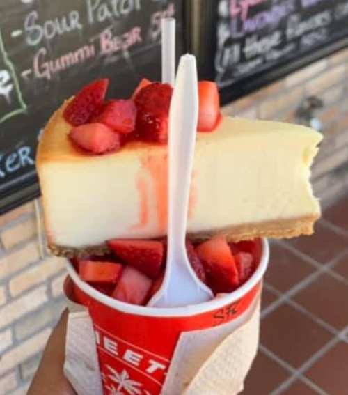 A slice of cheesecake topped with fresh strawberries, served in a red cup with a spoon, against a brick wall backdrop.