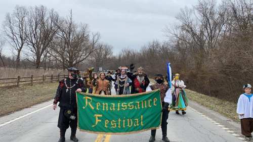 A group of people in Renaissance costumes parades down a road, holding a green banner for a Renaissance Festival.