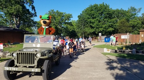 A large bear mascot sits in a military jeep, leading a group of children down a sunny path.