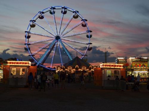 A vibrant carnival scene at sunset, featuring a Ferris wheel and colorful food stalls.