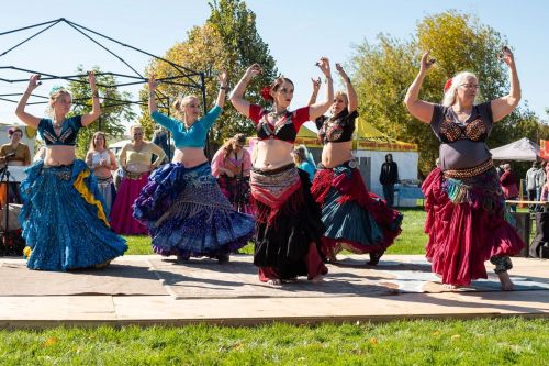 A group of dancers in colorful costumes perform a lively dance outdoors, surrounded by an audience and tents.