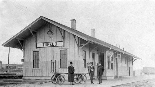Historic Tupelo train station with three people standing outside, featuring a wooden structure and a cart nearby.