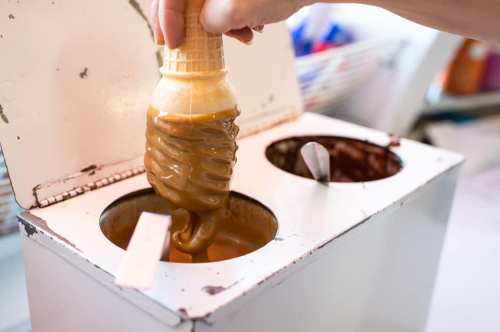A hand dips an ice cream cone into a container of caramel sauce, with another container visible nearby.