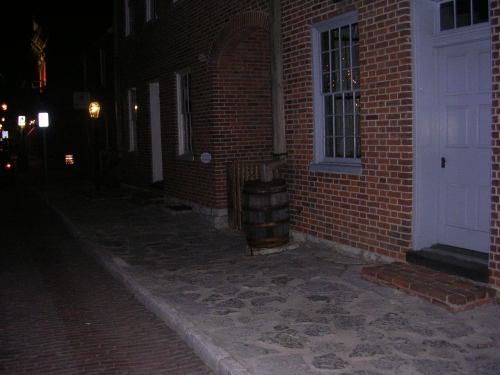 A dimly lit street at night featuring brick buildings, a wooden barrel, and a stone pathway.