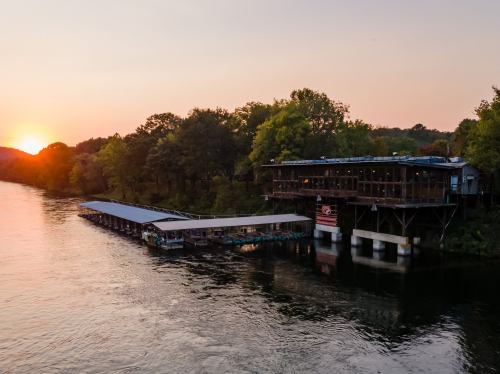 Sunset over a river with a wooden dock and a restaurant on stilts surrounded by trees.