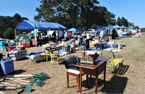 A bustling outdoor market with tents, various items for sale, and a mix of furniture and tools on display.