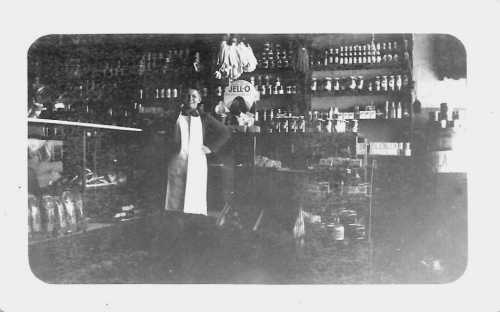 A vintage store interior with shelves of goods and two men, one in an apron, standing among the products.