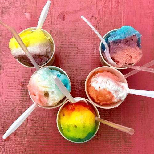 Five colorful cups of shaved ice in various flavors, each with a plastic spoon, arranged on a pink surface.