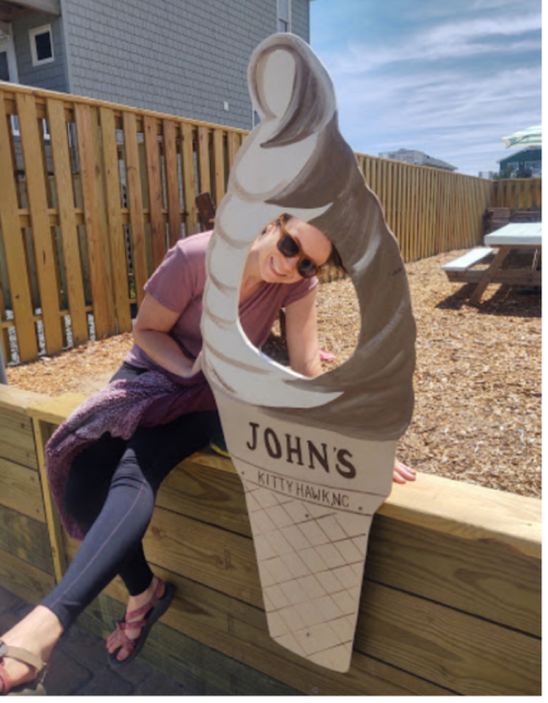 A person poses playfully with a large ice cream cone sign that reads "John's Kitty Hawk." Outdoor setting with wooden fence.