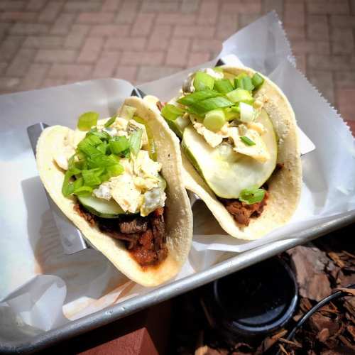 Two tacos filled with meat, topped with sliced green onions and a creamy sauce, served on a tray.