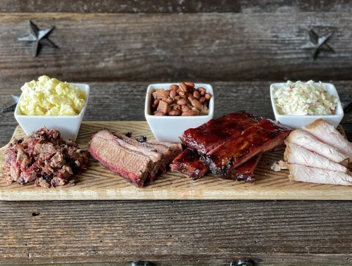 A wooden platter with various meats, including brisket, ribs, and pulled pork, alongside sides of coleslaw, beans, and potato salad.
