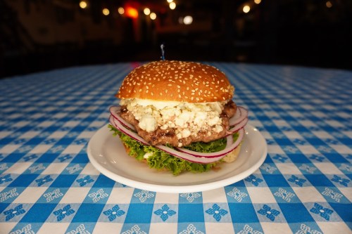 A sesame seed burger with lettuce, red onion, and a creamy topping, served on a white plate on a blue checkered tablecloth.