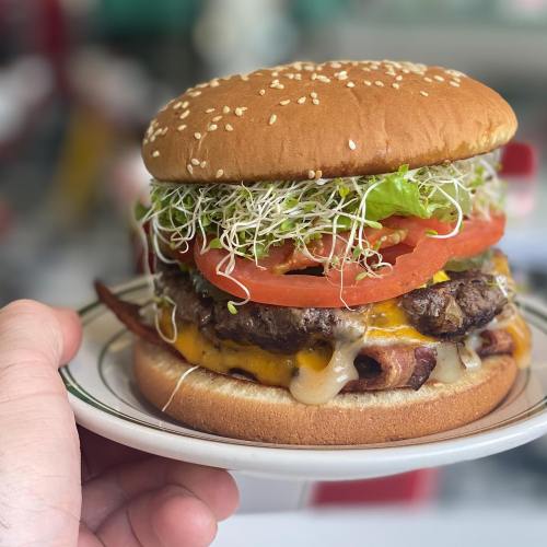 A hand holding a sesame seed bun burger stacked with lettuce, tomato, sprouts, cheese, and a juicy beef patty.
