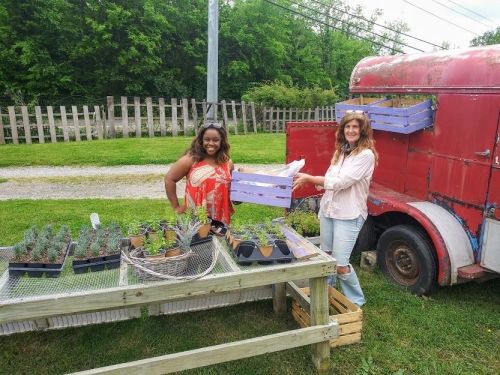 Two women smile while arranging plants on a table outside a vintage red trailer in a green garden setting.