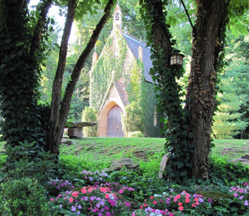 A quaint, ivy-covered chapel surrounded by trees and colorful flowers in a serene garden setting.