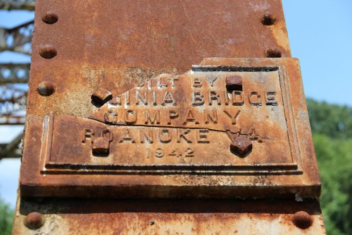 Close-up of a weathered metal plaque on a rusty bridge, reading "Built by Luna Bridge Company, Roanoke, VA, 1942."