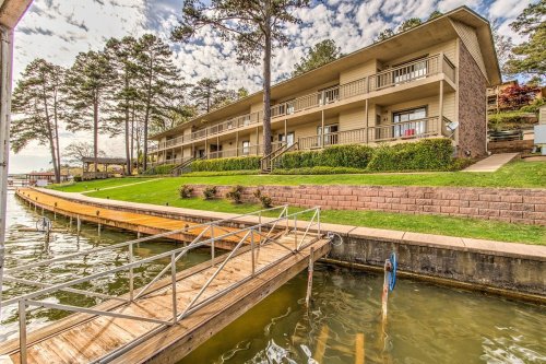 A lakeside building with balconies, surrounded by greenery and a wooden dock extending into the water.