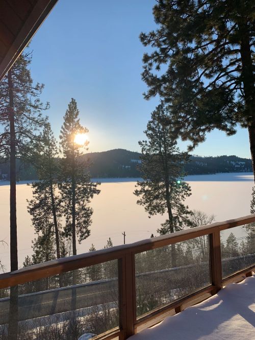 A serene lakeside view at sunrise, framed by tall pine trees and a wooden railing, with snow on the ground.
