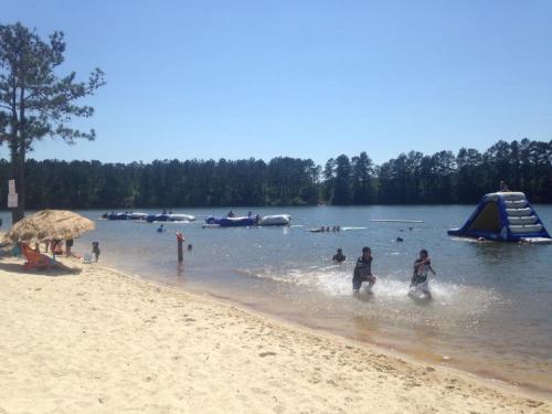 Two children splash in shallow water at a beach, with inflatable structures and people enjoying the lake in the background.
