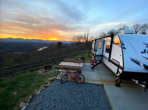 A camper parked on a scenic overlook at sunset, with a bike and picnic table nearby, surrounded by mountains and a river.
