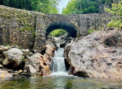 A stone bridge arches over a rocky stream, with water cascading down boulders into a serene pool below.
