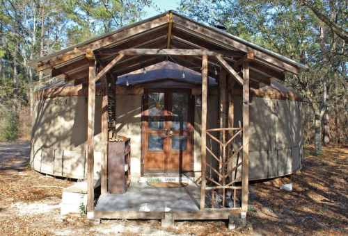 A round, wooden cabin with a porch, surrounded by trees and fallen leaves, in a natural outdoor setting.
