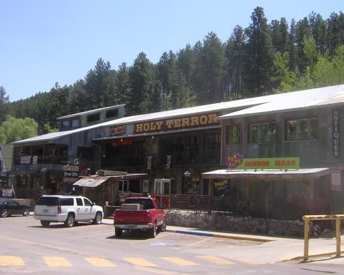 A rustic building with the sign "Holy Terror" and various shops, surrounded by trees and parked cars.