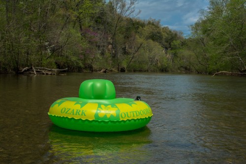 A bright green inflatable tube floats on a calm river surrounded by lush trees and a cloudy sky.