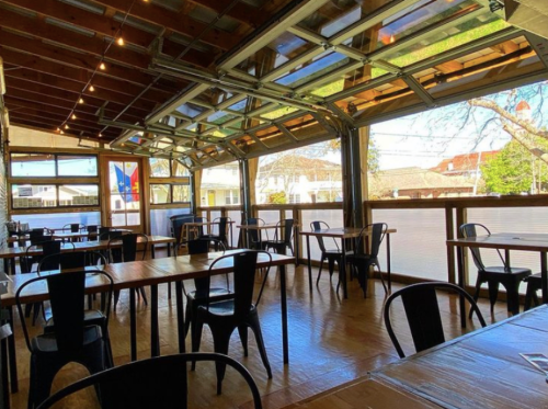 A spacious, modern restaurant interior with wooden tables and black chairs, featuring large open windows and string lights.