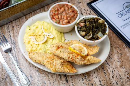 A plate of fried fish with lemon, served with mashed potatoes, collard greens, and a side of beans.