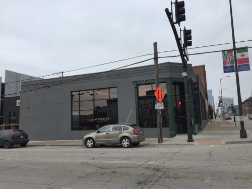 A gray building with large windows at a street corner, with a parked car and traffic signals in the foreground.