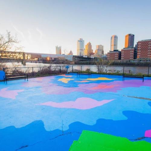 Colorful mural on a pavement with a city skyline and river in the background during sunset.