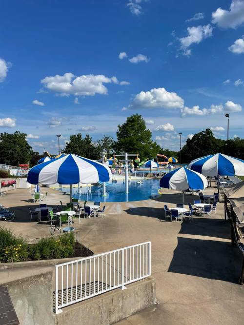 A sunny pool area with blue and white umbrellas, lounge chairs, and a clear sky filled with fluffy clouds.