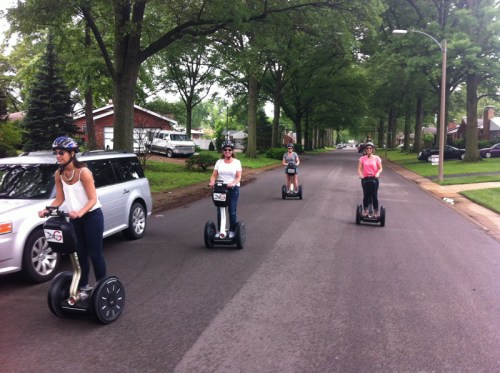Four people riding Segways down a tree-lined street, wearing helmets and casual clothing.