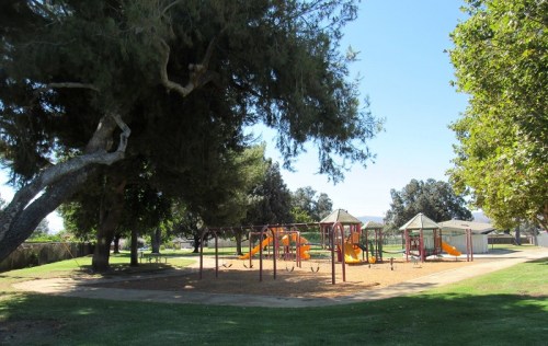 A sunny park featuring a playground with slides and climbing structures, surrounded by trees and grassy areas.
