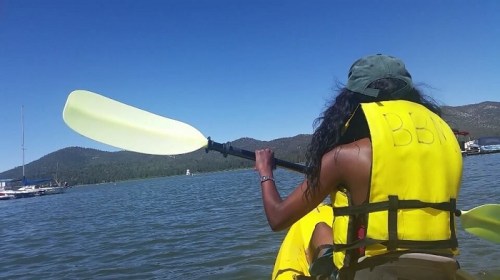 A person in a yellow life jacket paddles a kayak on a calm lake with mountains in the background.