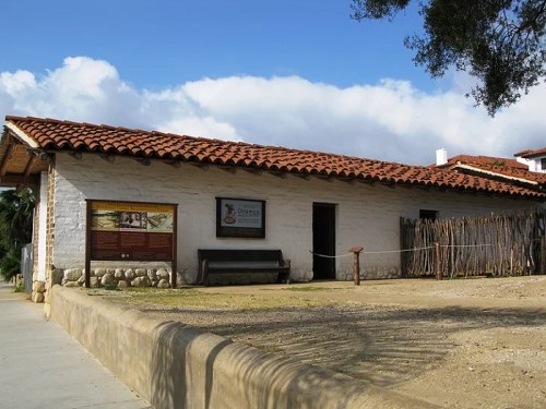 Historic building with a tiled roof, stone foundation, and informational signs, set against a blue sky with clouds.
