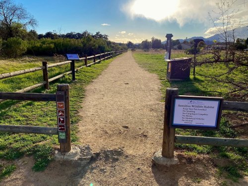 A dirt path through a green area, flanked by wooden fences and signs about wildlife habitat regulations.