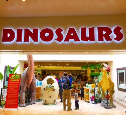 A family stands at the entrance of a dinosaur-themed attraction, featuring large dinosaur models and colorful decor.