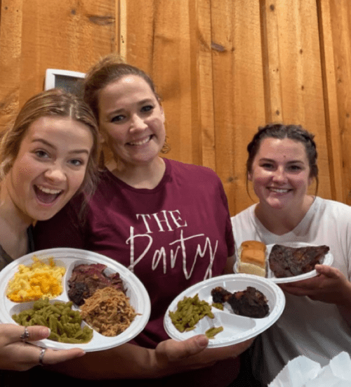 Three women smiling and holding plates of food, featuring meat, green beans, macaroni and cheese, and bread, in a cozy setting.