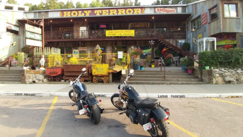Two motorcycles parked in front of a colorful antique shop named "Holy Terror" with vibrant decor and signage.