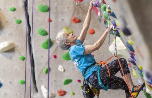 An older climber ascends a colorful indoor rock wall, focused and determined, with safety gear and harness.