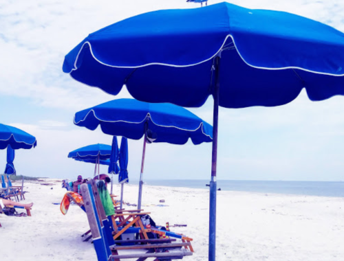 Blue beach umbrellas shade wooden lounge chairs on a sandy shore, with a calm sea in the background.