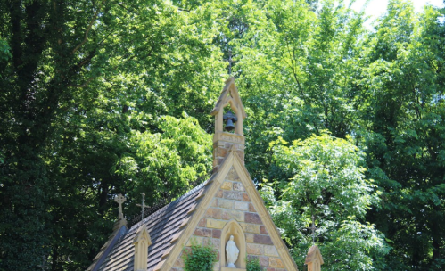 A stone chapel with a bell tower surrounded by lush green trees on a sunny day.