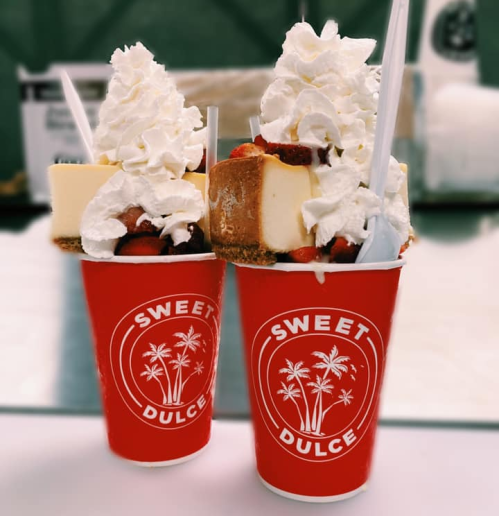 Two red cups filled with cheesecake topped with whipped cream and strawberries, labeled "Sweet Dulce."